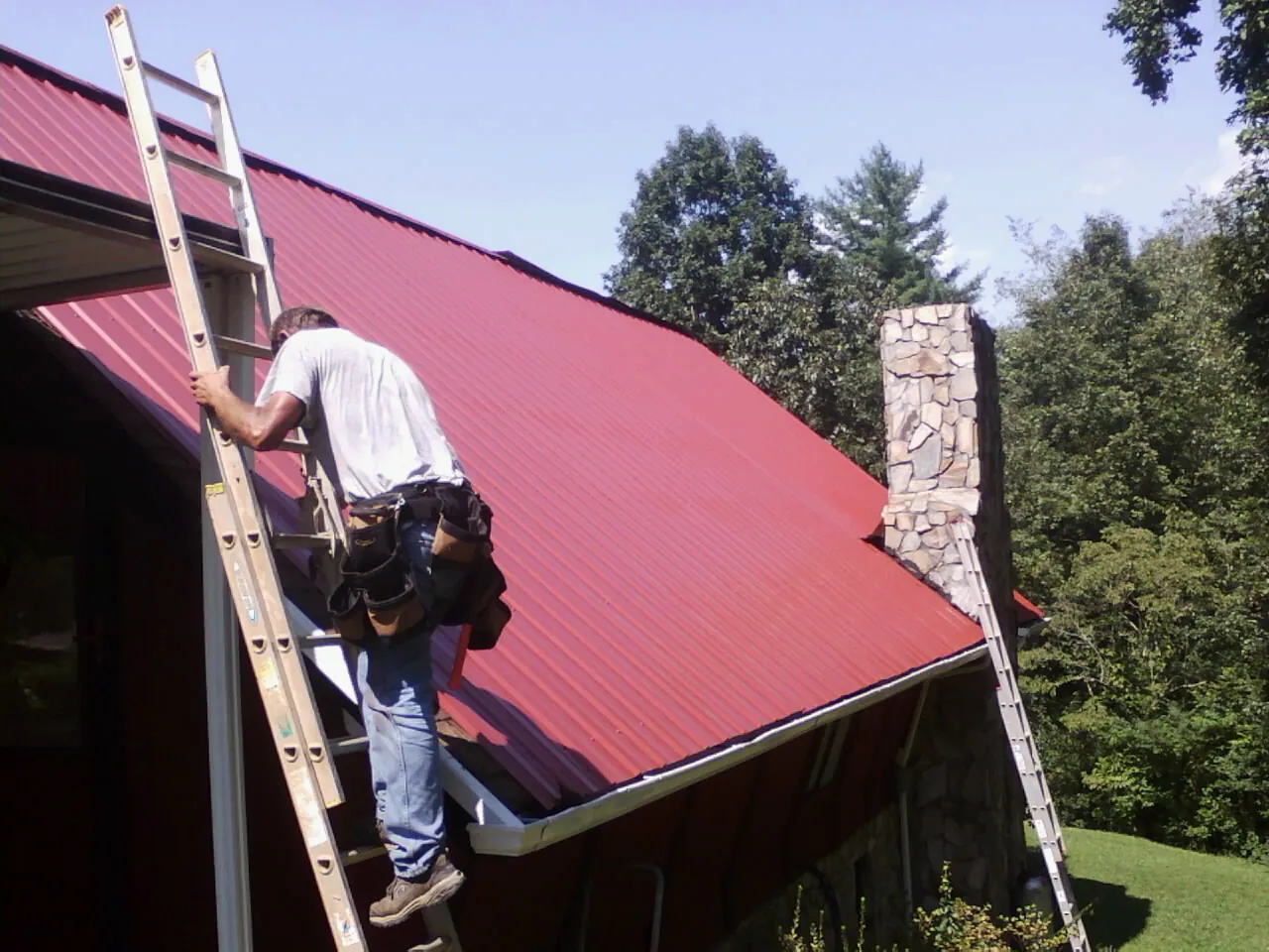 Roofer on a ladder beside a red metal roof and stone chimney — Soffit & Fascia Repair by Roofing Brothers in Nyssa